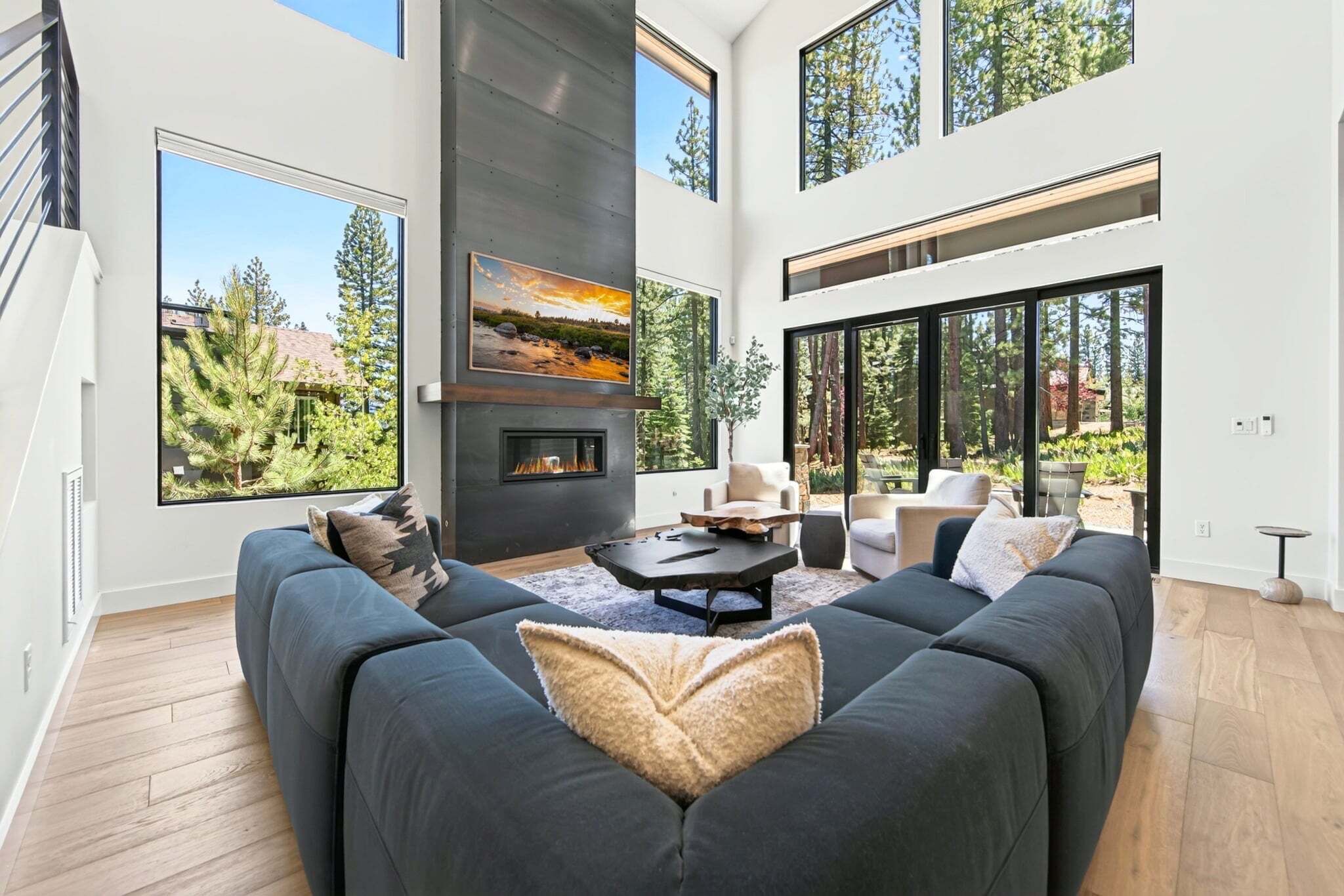 Una sala de estar moderna con un sofá grande, una chimenea y ventanas amplias que dan a un paisaje de árboles.