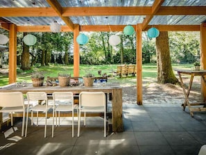 An outdoor covered patio with a wooden table and chairs, surrounded by trees and greenery.