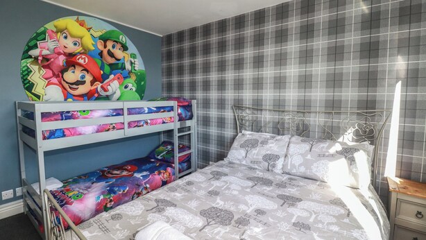 A bedroom with a bunk bed, a bed with patterned bedding, a wall clock, and a window with a view. - Shoreline Apartment No.8 (Newquay)