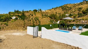 Ein Poolbereich mit weißer Wand, einem Tor und Blick auf einen Hang mit Häusern.