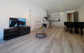 A modern living room with a flat-screen TV, a dining table with chairs, a small round table with a plant, and a kitchen area with cabinets and appliances.
