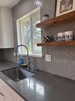 A kitchen with a herringbone tile backsplash, a stainless steel sink, and wooden shelves with mugs. - Lovely updated 3-bedroom house in quaint Cortez perfect for your getaway (Cortez)