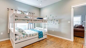 A room with a bunk bed, wooden flooring, a wall with decorative decals, and a brown leather sofa in the adjacent room.