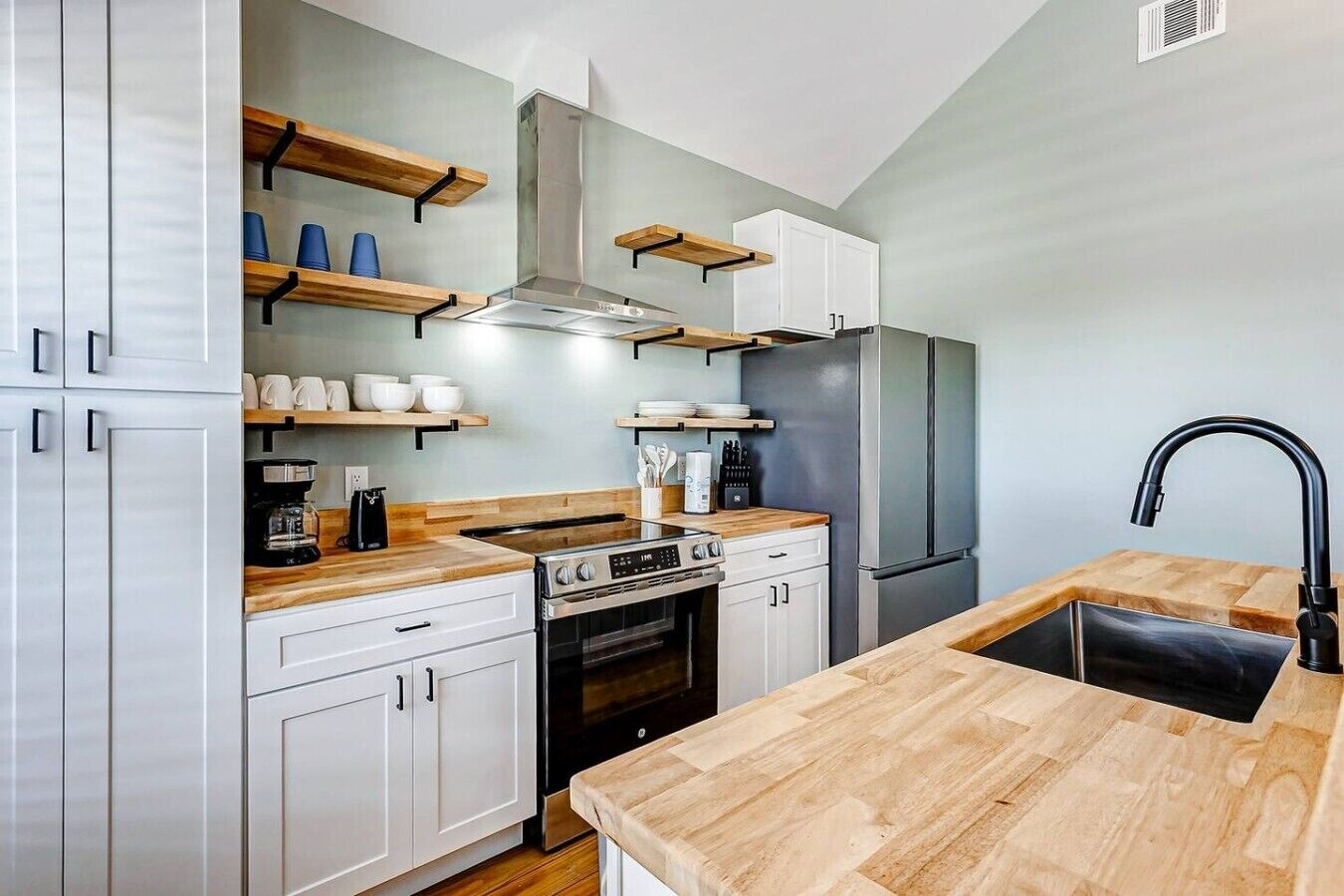 Una cocina moderna con armarios blancos, encimeras de madera, extractor de acero inoxidable, horno empotrado, refrigerador y grifo negro.