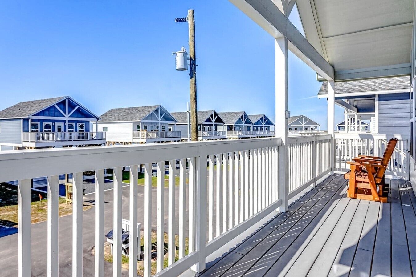 A wooden deck with a white railing, a wooden chair, and a view of beach houses.