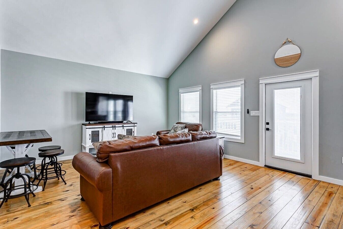 A living room with a brown leather sofa, a TV on a cabinet, and a dining table with stools.