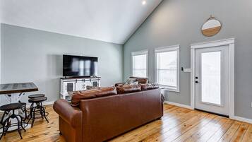A living room with a brown leather sofa, a TV on a cabinet, and a dining table with stools.