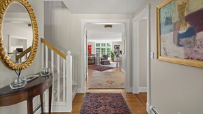 Ein Flur mit einer Holztreppe, einem großen Spiegel, einem Essbereich mit Tisch und Stühlen sowie einem Gemälde an der Wand.
