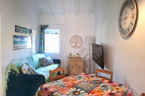 Ein Zimmer mit einem Bett, einem Tisch mit blumigem Tischläufer, einer Uhr und Bildern an der Wand.