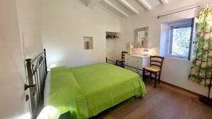 Ein Schlafzimmer mit Bett, Schminktisch, Stuhl, Fenster mit Vorhängen und Spiegel an der Wand.
