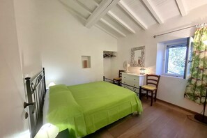 Ein Schlafzimmer mit Bett, Schminktisch, Stuhl, Fenster mit Vorhängen und Spiegel an der Wand.