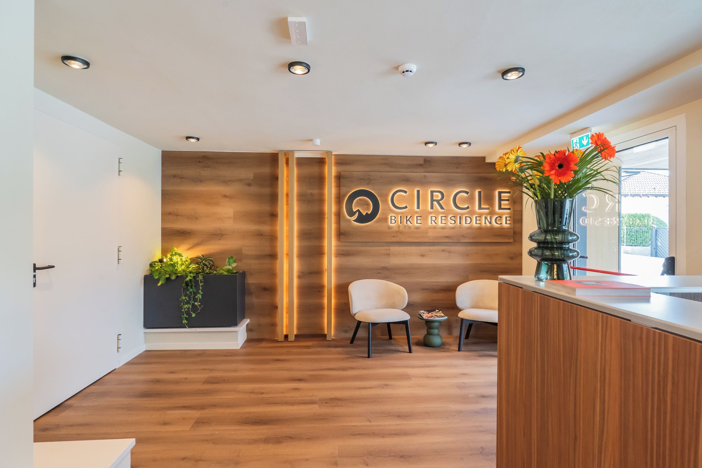 Ein Empfangsbereich mit einer Holzwand, einem Schild mit der Aufschrift 'Circle Bike Residence', zwei Stühlen, einer Pflanze und einer Vase mit Blumen.