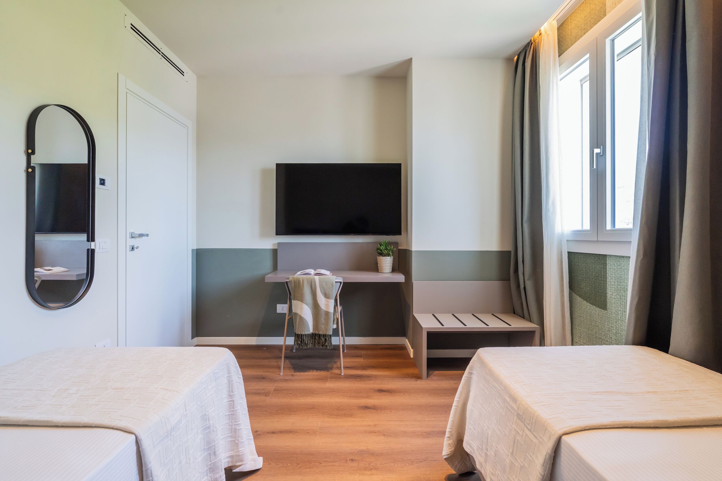 Una camera d'albergo con due letti, una scrivania con una sedia, una televisione e una finestra con tende.