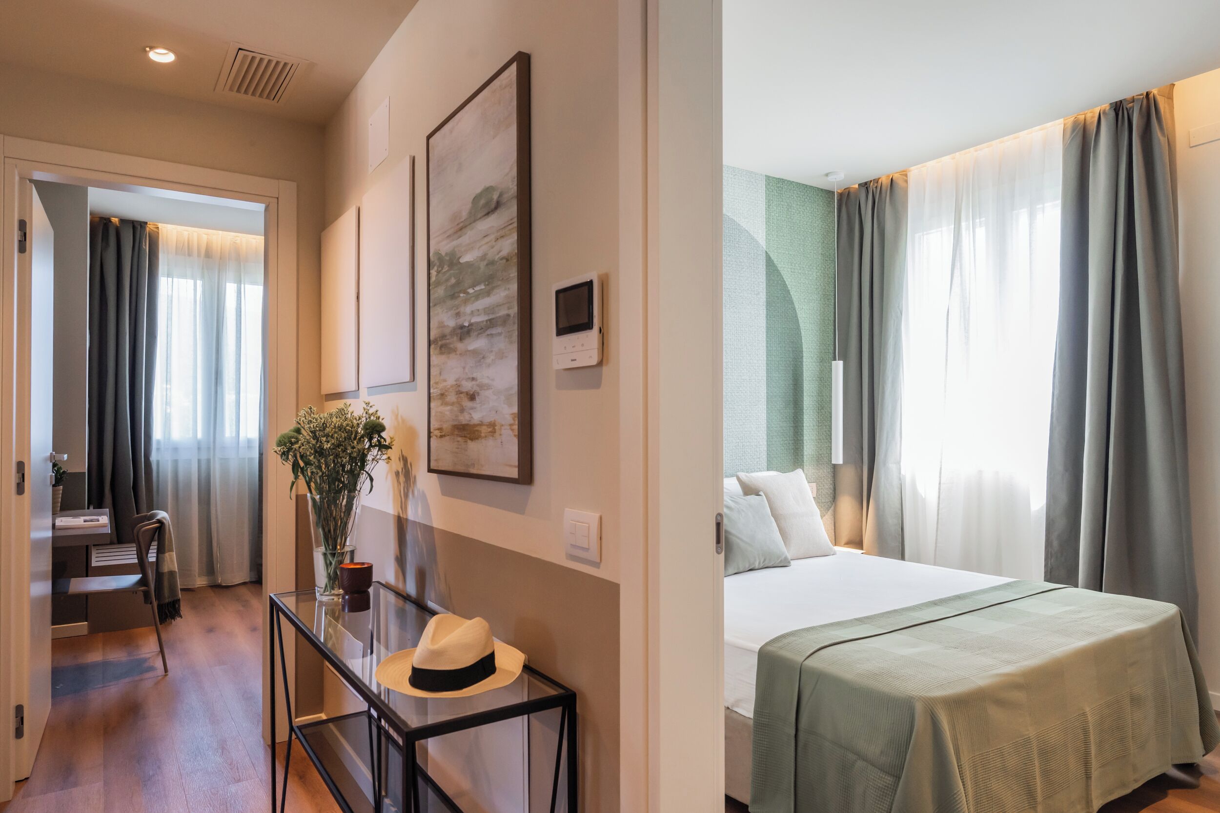Quarto de hotel com cama, escrivaninha com chapéu, vaso com flores e um quadro na parede.