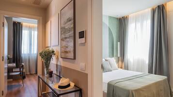 Quarto de hotel com cama, escrivaninha com chapéu, vaso com flores e um quadro na parede.