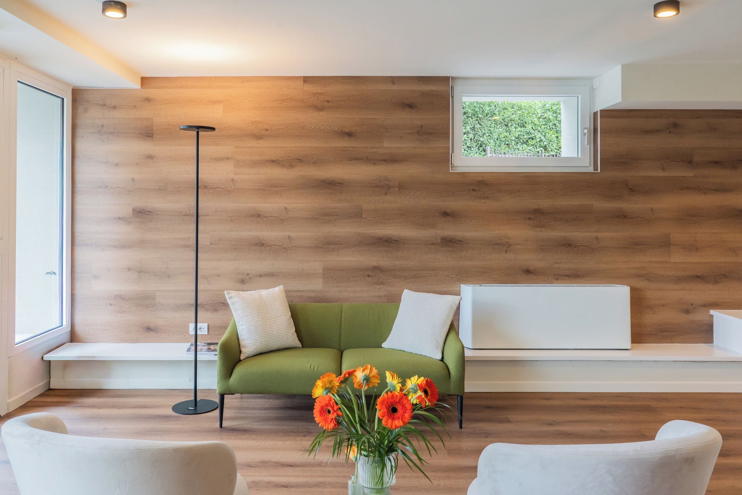 Ein modernes Wohnzimmer mit einer Holzwand, einem grünen Sofa mit Kissen, einer Stehlampe und einer Vase mit Blumen.