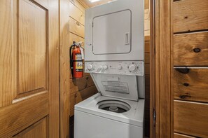 A compact laundry room with a washing machine and dryer stacked vertically, wooden walls, and a fire extinguisher mounted on the wall. - Mountain Movie Night in Gatlinburg (Gatlinburg)