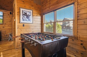 Habitación con piso de madera, una mesa de fútbol de mesa, una fotografía enmarcada en la pared y un ventanal con persianas.