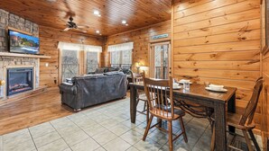 Interior acogedor de una cabaña con chimenea, un sofá, una mesa de comedor con sillas y un televisor montado en la pared.