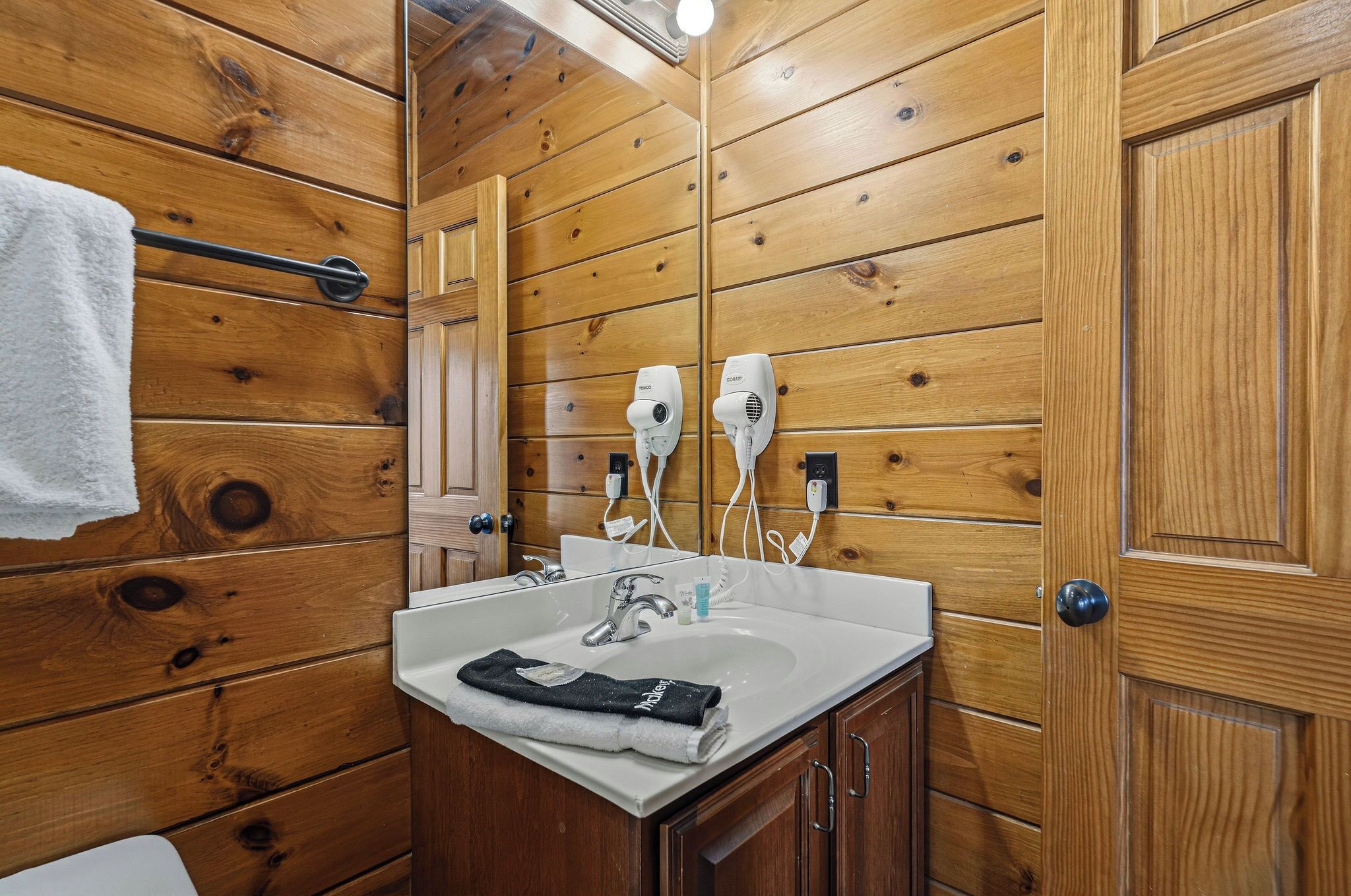 Een badkamer met houten lambrisering, een wastafel, een handdoekhouder en twee aangesloten haardroger.