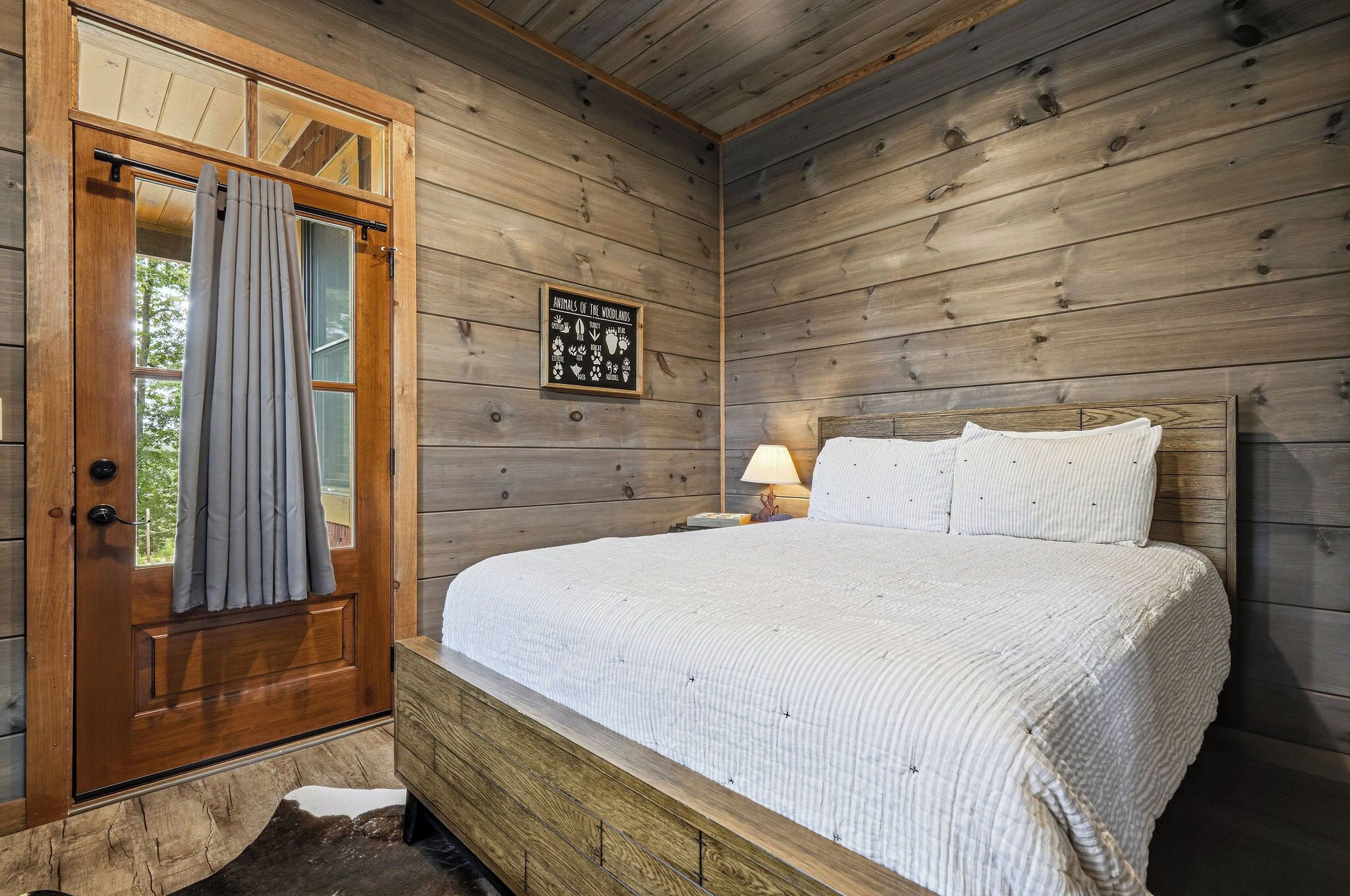 Ein Zimmer aus Holz mit einem Bett, einem Nachttisch, einer Lampe und einer Tür mit Vorhängen.