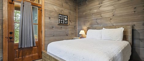 Una habitación de madera con cama, mesita de noche, lámpara y una puerta con cortinas.