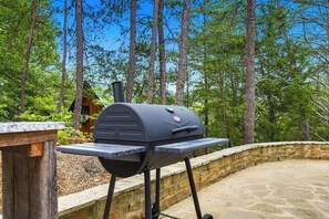 A barbecue grill with a stone wall and wooden deck, set against a backdrop of tall trees and a clear sky. - Modern Rustic Charm in Pigeon Forge (Sevierville)