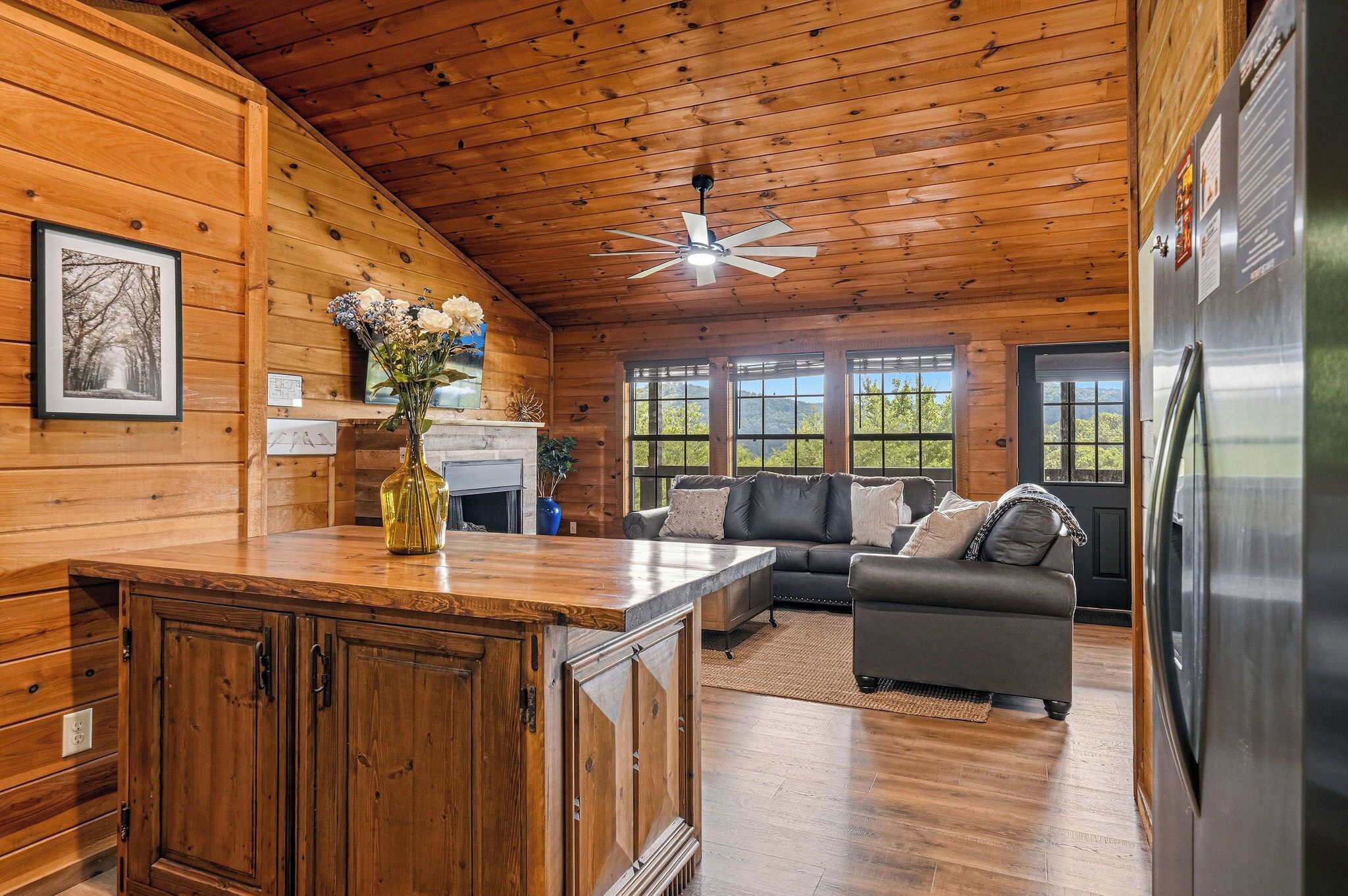 A cabin interior with a wooden ceiling, a ceiling fan, a fireplace, a sofa, and a refrigerator.