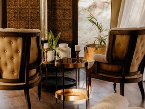 A room with two tufted armchairs, a small round table with a candle, and a wicker basket with plants.