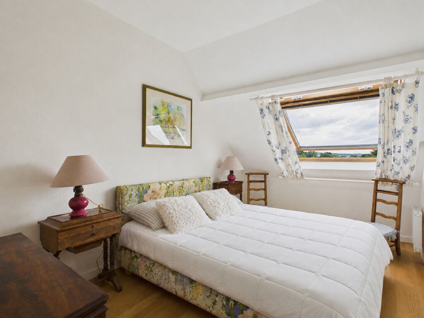 A bedroom with a bed, two bedside tables, a window with curtains, a wooden floor, and a wall-mounted picture.