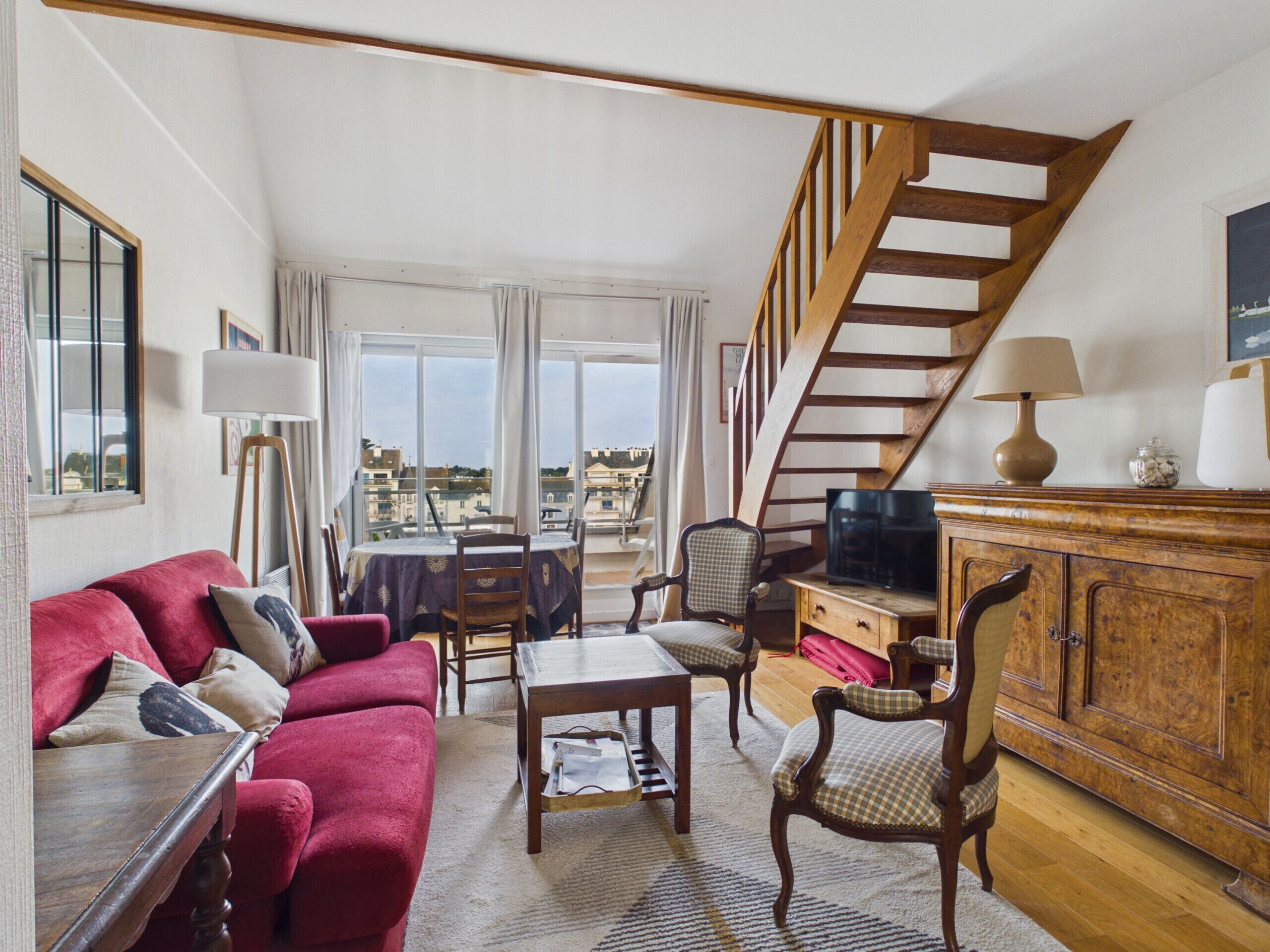 A spacious living area with a red sofa, a wooden coffee table, and a television. A staircase with wooden railings leads to an upper floor.