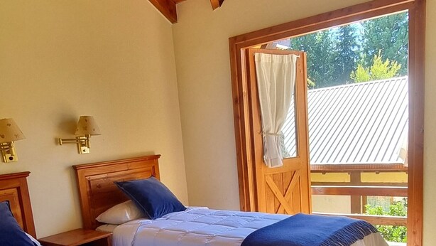 A room with two beds, wooden beams, a window with curtains, and a view of greenery outside.