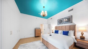 Une chambre moderne avec un plafond de couleur turquoise, un grand lit agrémenté de plusieurs coussins, une commode en bois et une table de chevet avec une lampe.