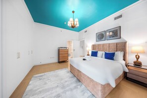 Ein modernes Schlafzimmer mit himmelblau gestrichener Decke, einem großen Bett mit vielen Kissen, einer Holzkommode und einem Nachttisch mit Lampe.