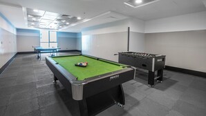 Une table de billard recouverte d’un feutre vert et une table de baby-foot se trouvent dans une pièce aux murs blancs et au sol carrelé.