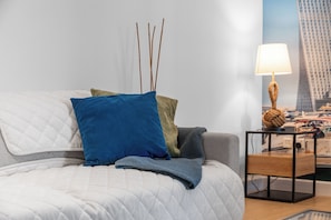 Un lit recouvert d’une couette blanche matelassée, un oreiller bleu et une couverture grise. Sur une table de chevet en bois, à côté du lit, se trouve une lampe dont le pied est fait de corde.