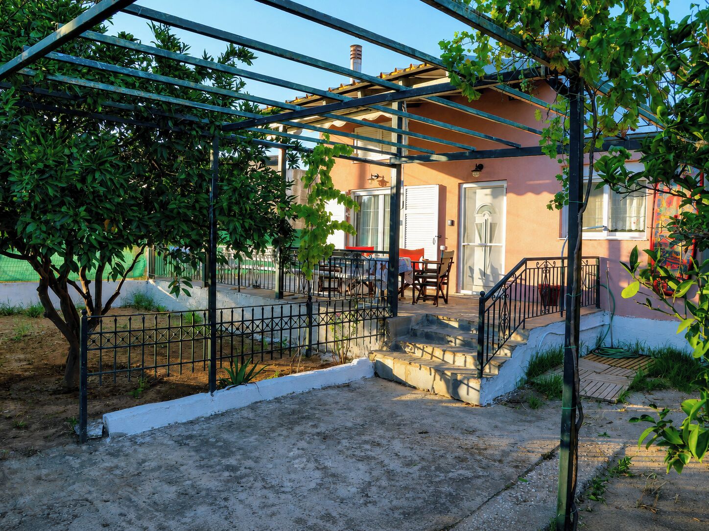 Ett takförsett utomhusområde med en pergola av metall, en trappa som leder till en byggnadsingång samt ett litet bord och några stolar.