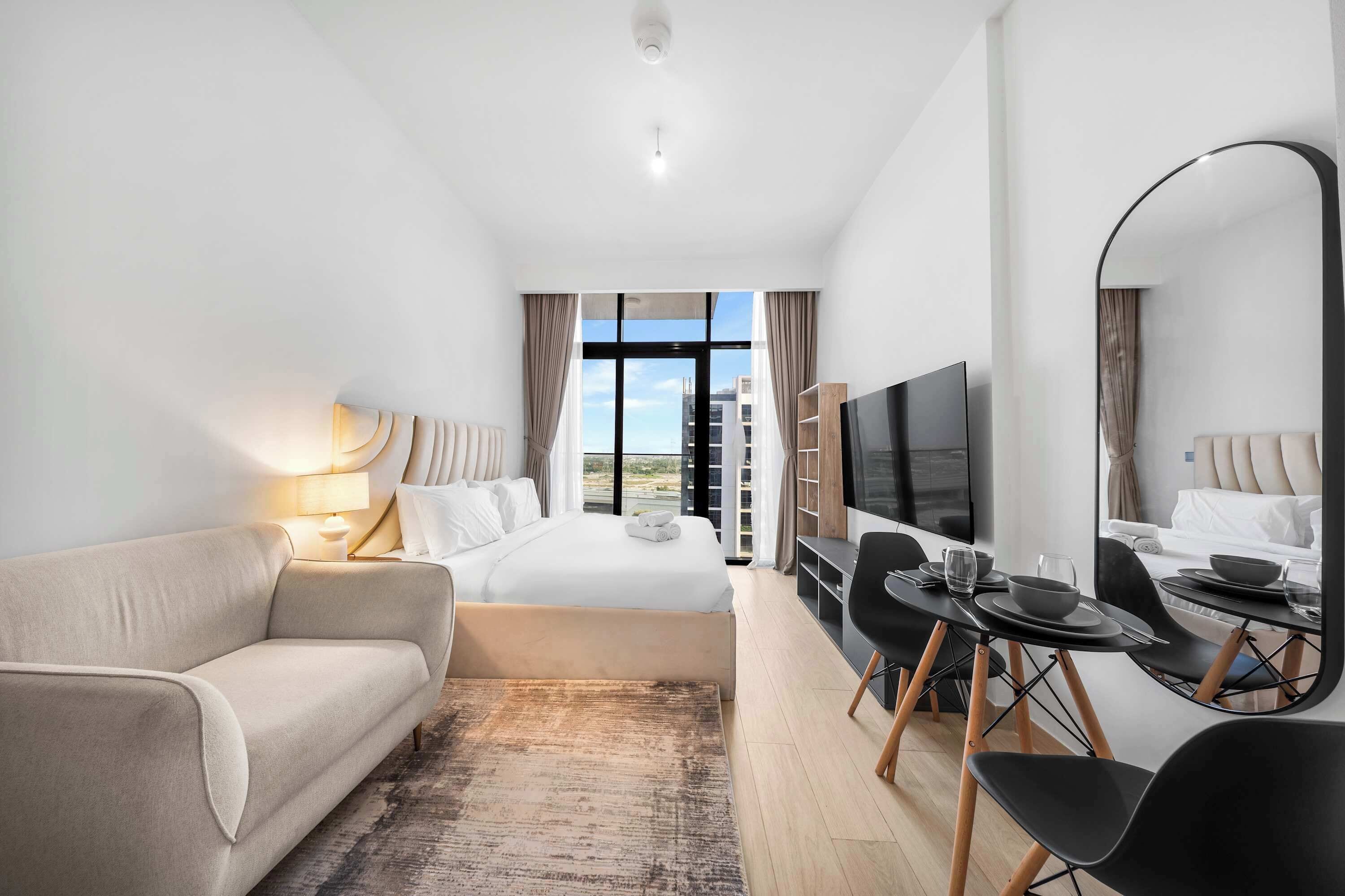 A hotel room with a bed, a sofa, a TV, a dining table with chairs, and a large window with a view.