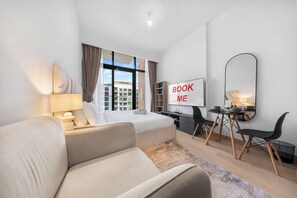 A modern hotel room with a bed, a desk, a chair, a sofa, and a large window. - StayShort - Luxury Studio Apartment in Meydan with Balcony (Meydan)