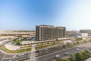 A modern multi-story building with a flat roof, surrounded by a landscaped area with palm trees and a road with cars. - StayShort - Luxury Studio Apartment in Meydan with Balcony (Meydan)