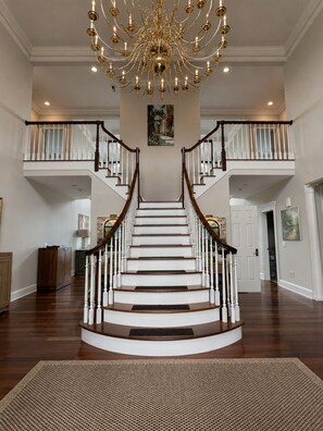 A grand staircase with a chandelier, white railings, and a rug at the bottom.