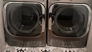 A laundry area with two front-loading washing machines, a cabinet above, and a water softener unit to the right.