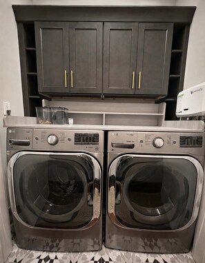 A laundry area with two front-loading washing machines, a cabinet above, and a water softener unit to the right.