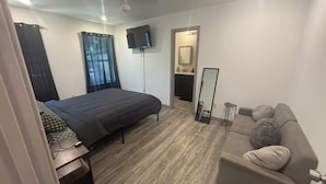 A bedroom with a bed, a sofa, a TV, a mirror, and a bathroom visible through an open door. - Herrling Home (Dallas)