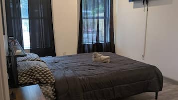 Un dormitorio con cama, escritorio, silla, televisor montado en la pared y dos ventanas con cortinas.