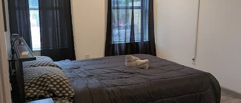 A bedroom with a bed, a desk, a chair, a TV mounted on the wall, and two windows with curtains.