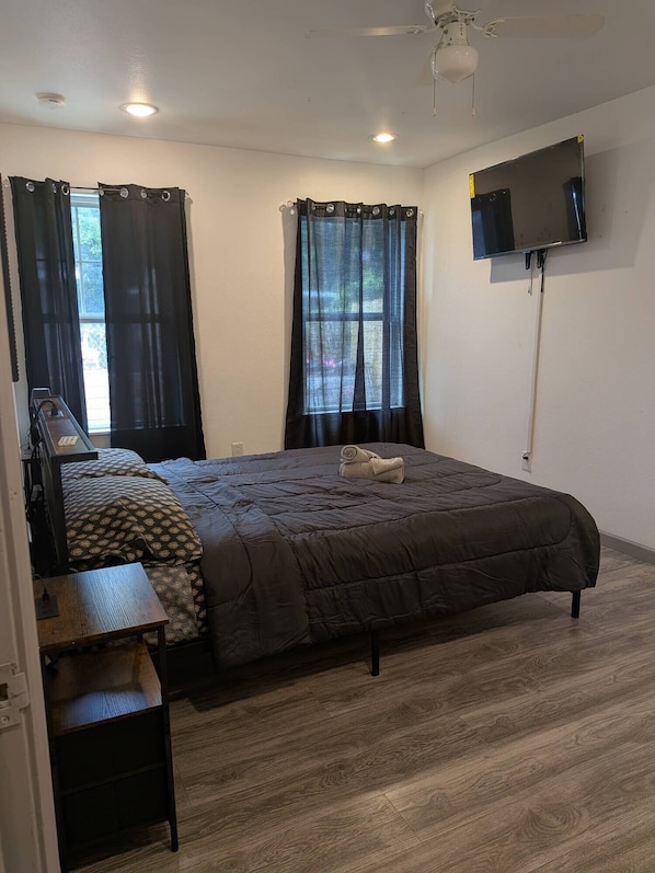 A bedroom with a bed, a desk, a chair, a TV mounted on the wall, and two windows with curtains.