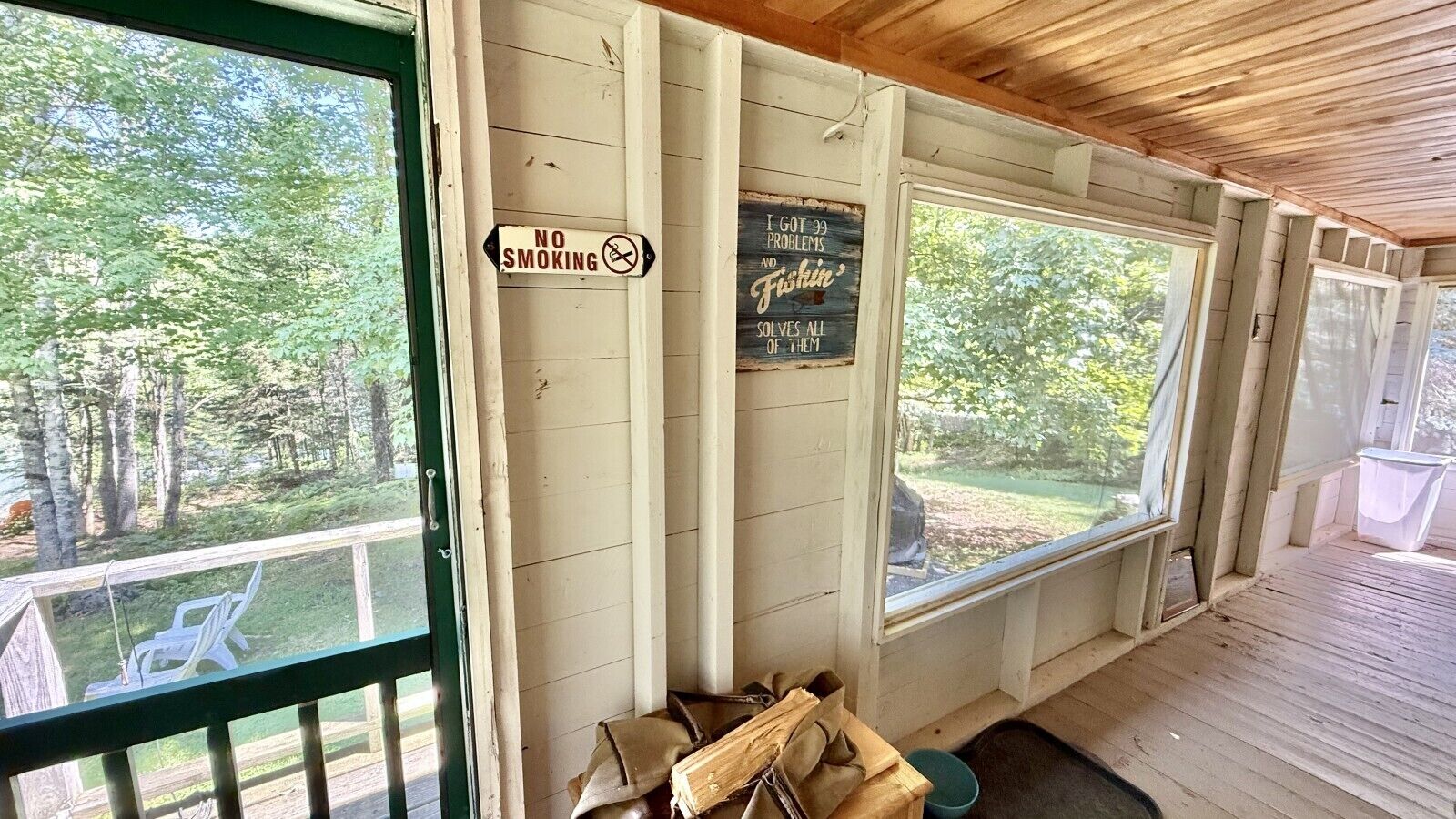 Eine überdachte Veranda mit einem 'RAUCHVERBOT'-Schild, einem Schild mit der Aufschrift 'ICH HABE 99 PROBLEME, ABER ANGELN LÖST ALLE' und Blick auf ein bewaldetes Gebiet.