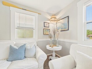 A room with a white sofa, a small round table with decorative items, a framed picture on the wall, and a window with blinds.