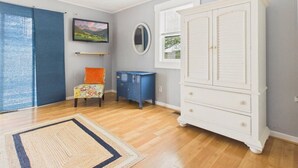Una habitación con suelo de madera, un armario blanco, un aparador azul, una silla colorida y un ventanal con persianas.